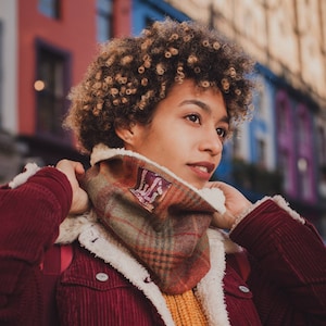 May include: A woman with curly brown hair is wearing a red corduroy jacket with a brown and green plaid scarf. The scarf has a white fleece lining and a small patch with a design on it.