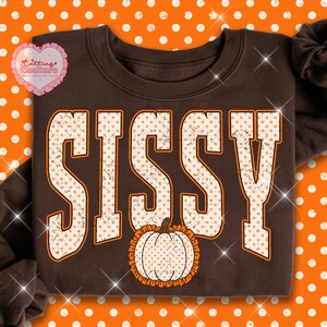 May include: A dark brown crewneck sweatshirt featuring the word SISSY in large, block-style letters with a white and orange polka dot pattern. Below the text is a white pumpkin graphic with an orange ruffled border, all set against a brown fabric.