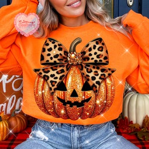 May include: An orange long-sleeve sweatshirt featuring a large graphic of a glittery orange jack-o'-lantern pumpkin topped with a prominent leopard print bow. The pumpkin has black triangular eyes and a jagged black mouth design.