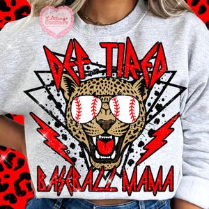 May include: A light gray sweatshirt with the text "DF TIRED BASEBALL MAMA" in red. The design features a leopard wearing baseball-themed sunglasses, with red lightning bolts and geometric shapes.