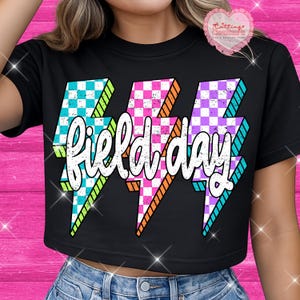 May include: Black crop top with three lightning bolt graphics in teal, pink, and purple checkered patterns. The word "field day" is written in white script across the bolts. The shirt is worn with blue jeans.