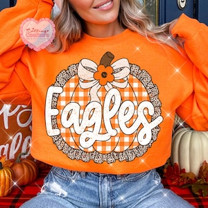 May include: A bright orange long-sleeve sweatshirt featuring a large graphic of a pumpkin with a white and orange checkered pattern. The pumpkin is accented with a white bow, a small orange flower, and a leopard print border. The word Eagles is printed.