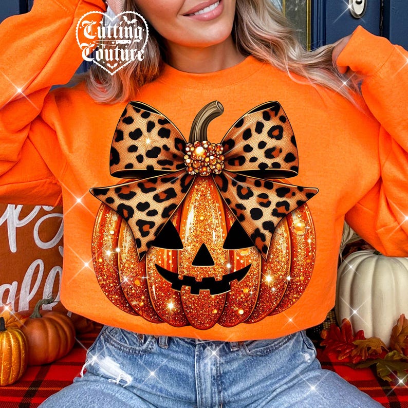 May include: An orange long-sleeve sweatshirt featuring a graphic of a glittery orange jack-o'-lantern pumpkin topped with a large leopard print bow. The design includes the text Cutting Couture in the upper left corner of the graphic.