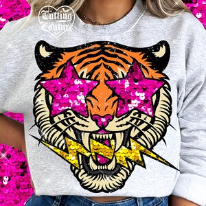 May include: A light gray sweatshirt featuring a tiger face with star-shaped sequined sunglasses and a gold lightning bolt. The tiger has orange and black stripes, with a pink nose and mouth. The text "Cutting Couture" is at the top.