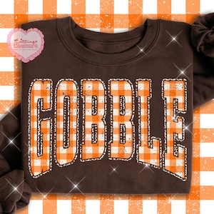 May include: A dark brown crewneck sweatshirt featuring the word GOBBLE in large, distressed, orange and white chequered block letters. The text has a textured, worn appearance and is centred on the front of the garment.
