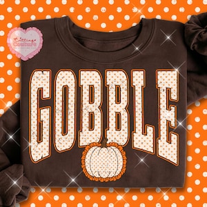 May include: A dark brown crewneck sweatshirt featuring the word GOBBLE in large, block-style letters with a distressed, light orange polka dot pattern. Below the text is a graphic of a pumpkin with a matching orange polka dot design.