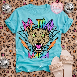 May include: A light blue t-shirt with a leopard graphic and the words "AF TIRED MAMA" in a colorful, retro font. The leopard has green eyes and an open mouth. The shirt is displayed with white sandals.