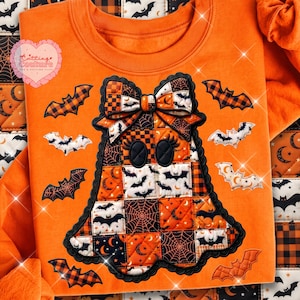 May include: An orange sweatshirt featuring a large, quilted ghost applique with a bow on its head. The ghost is constructed from patchwork squares displaying Halloween patterns like bats, moons, and spiderwebs. Several individual bat appliques surround the central ghost figure.