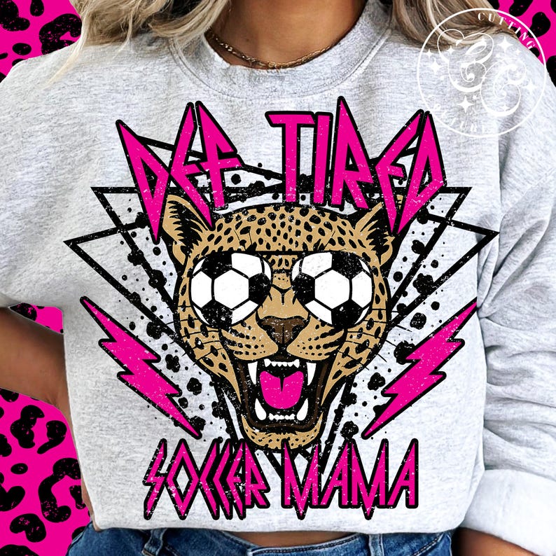 K&ouml;nnte beinhalten: Ein graues Sweatshirt mit einem pinkfarbenen und schwarzen Grafikdesign, das einen Geparden mit Fu&szlig;b&auml;llen als Augen zeigt. Der Text "DEF TIRED SOCCER MAMA" ist in Pink gedruckt.