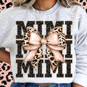 Pu&ograve; includere: Una felpa grigia con la parola "MIMI" stampata in lettere nere con un motivo leopardato. Un grande fiocco rosa con dettagli di glitter dorati &egrave; centrato tra le due parole.