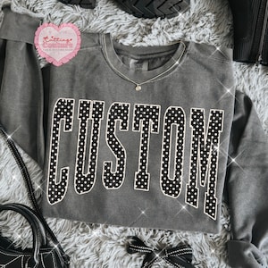 Peut inclure: Un sweat-shirt gris avec le mot "CUSTOM" en grandes lettres blanches à pois. Un petit collier argenté avec un pendentif est visible. Le sweat-shirt fait partie d'une collection de vêtements.