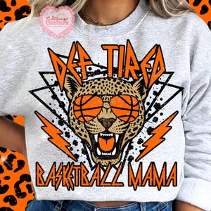 May include: A light grey sweatshirt with a graphic design featuring a leopard wearing basketball sunglasses. The text reads "Off Tired Basketball Mama" in orange and black.