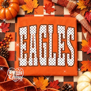 May include: An orange t-shirt featuring the word EAGLES in large, bold, block letters. The letters are white with a black polka dot pattern and a dark brown outline. The shirt is displayed on a background of orange and brown plaid fabric.