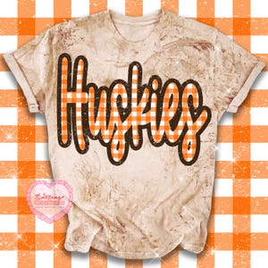 May include: A short-sleeved t-shirt with a bleached, mottled beige finish. The word HUSKIES is printed across the chest in a bold, dark brown outlined font filled with an orange and white gingham check pattern.