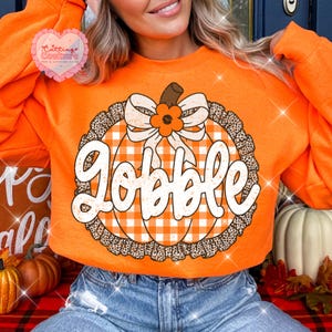 May include: An orange long-sleeve sweatshirt featuring a central graphic of a pumpkin with a white and orange gingham pattern, a leopard print border, and a white bow with an orange flower. The word Gobble is printed in white script across the pumpkin.