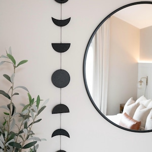 May include: A black metal moon phase wall hanging with a chain connecting each crescent moon shape. The hanging is on a white wall with a round mirror reflecting a room.