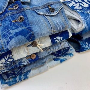May include: A stack of blue denim jackets with white floral patterns. The jackets are folded neatly and have a distressed look.