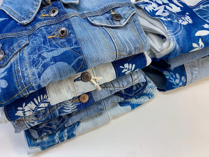 May include: A stack of blue denim jackets with white floral patterns. The jackets are folded neatly and have a distressed look.