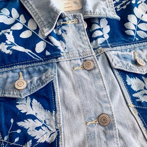 May include: A light blue denim jacket with white floral designs. The jacket has a button closure and two pockets.