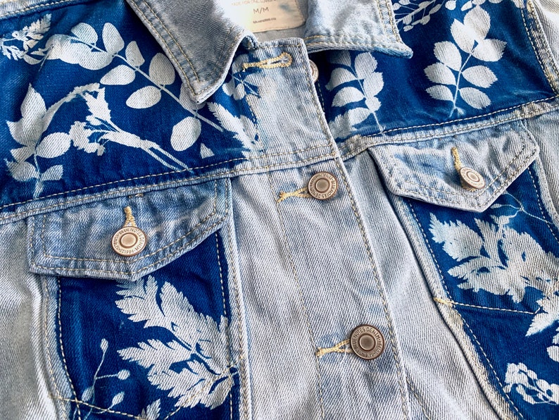 May include: A light blue denim jacket with white floral designs. The jacket has a button closure and two pockets.