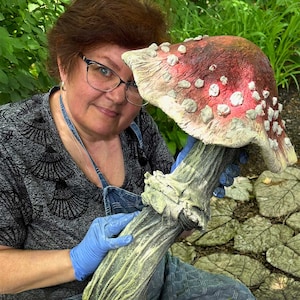 May include: A woman wearing blue gloves holds a large, hand-painted, red and white mushroom sculpture. The mushroom has a textured surface and white spots. The sculpture is made of concrete and has a realistic appearance.