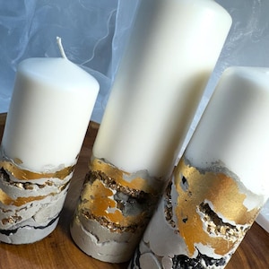 Könnte beinhalten: Drei weiße Kerzen in Säulenform mit einer dekorativen Schicht aus grauem Beton und goldenen Blattverzierungen. Die Kerzen sind in abnehmender Größe von links nach rechts angeordnet.