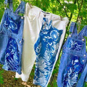 May include: Three pairs of denim overalls hanging on a clothesline outdoors. The overalls are decorated with white botanical prints, including ferns and dandelions. One pair of overalls is white denim.