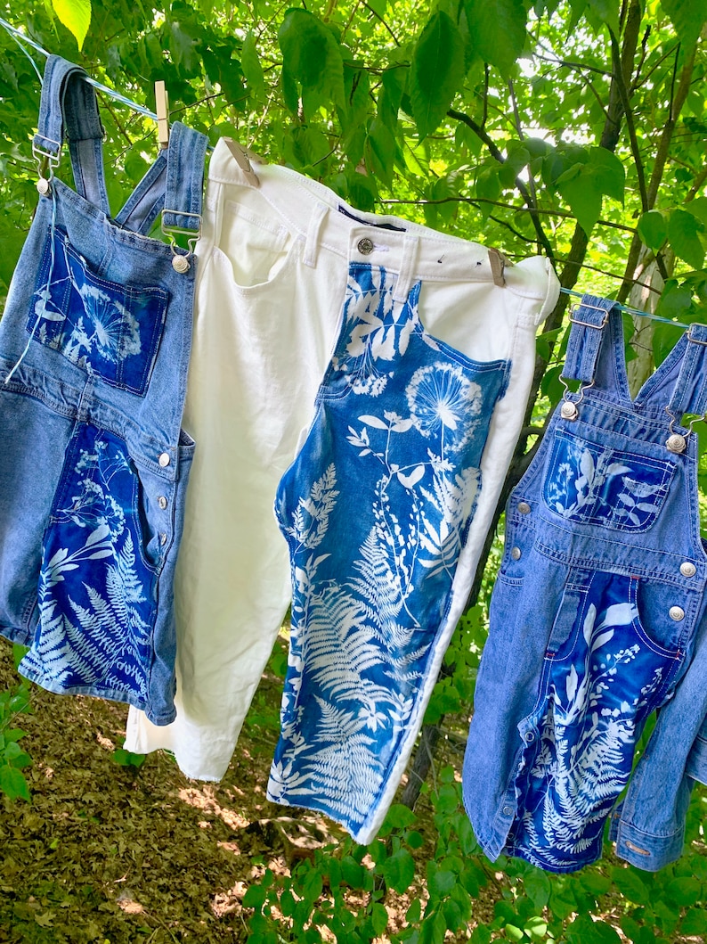 May include: Three pairs of denim overalls hanging on a clothesline outdoors. The overalls are decorated with white botanical prints, including ferns and dandelions. One pair of overalls is white denim.