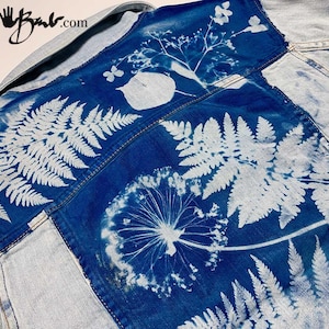 May include: A blue denim jacket with a white floral design. The design includes ferns, dandelions, and other botanical elements. The text "made by" and a handprint are visible on the top left corner of the jacket.