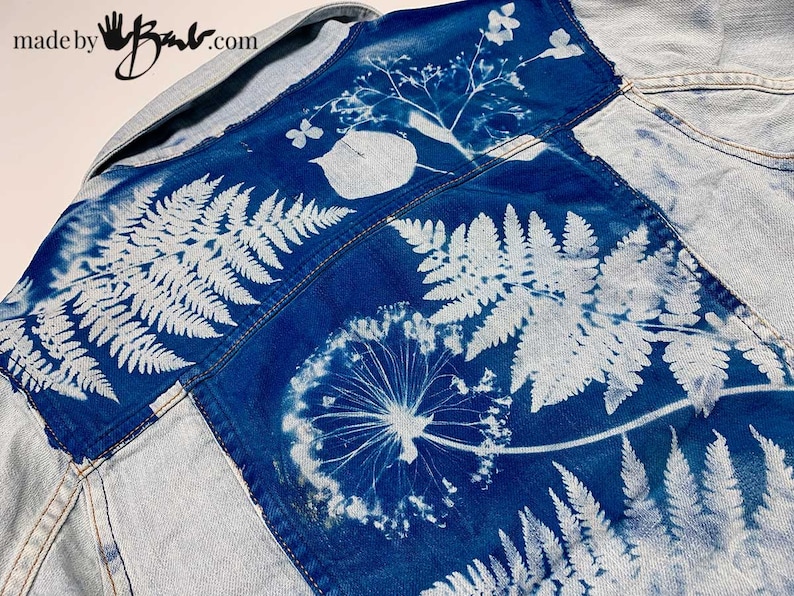 May include: A blue denim jacket with a white floral design. The design includes ferns, dandelions, and other botanical elements. The text "made by" and a handprint are visible on the top left corner of the jacket.