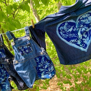 May include: Two pairs of denim overalls with white floral designs and a denim shirt with a white heart-shaped floral design. The clothing is hanging on a clothesline outdoors.