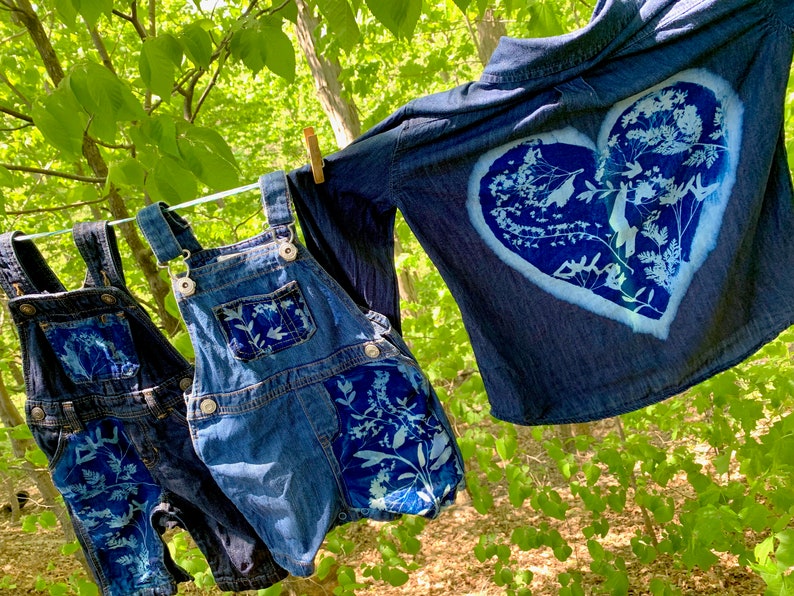 May include: Two pairs of denim overalls with white floral designs and a denim shirt with a white heart-shaped floral design. The clothing is hanging on a clothesline outdoors.