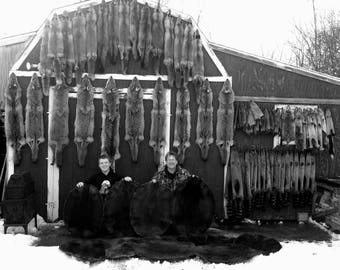May include: Two people stand in front of a wall lined with dozens of animal pelts. The pelts are mostly gray and brown, and some are hanging from the ceiling. The people are wearing casual clothing and are smiling. There are three large, round, dark-colored rugs on the floor in front of them.