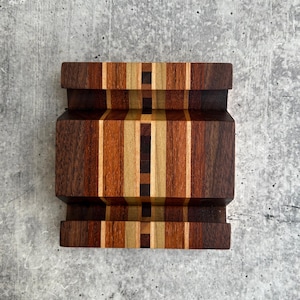 May include: A rectangular wooden phone stand featuring a multi-tonal striped pattern. The stand is constructed from various wood species in shades of dark brown, medium brown, tan, and cream, with a central vertical mosaic of small squares.
