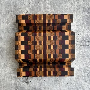 May include: A rectangular wooden stand featuring a complex mosaic pattern composed of small, square blocks in various shades of brown, tan, dark purple, and black. The object has a tiered design with two horizontal grooves carved into the surface.