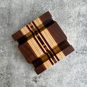May include: A wooden phone stand featuring a multi-colored striped pattern created from various wood species. The design includes alternating bands of dark brown, light tan, and deep purple wood, arranged in a geometric, layered construction.