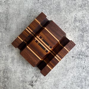 May include: A wooden phone stand featuring a dark brown walnut base with inlaid light and dark wood stripes. The stand has a stepped, tiered design with a central groove and two side sections, showcasing a geometric pattern of contrasting wood grains.