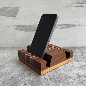 May include: A dark gray smartphone resting in a rectangular wooden stand. The stand is crafted from multiple wood tones, featuring a light brown base, a dark brown middle section, and inlaid geometric patterns in red, white, and dark wood.