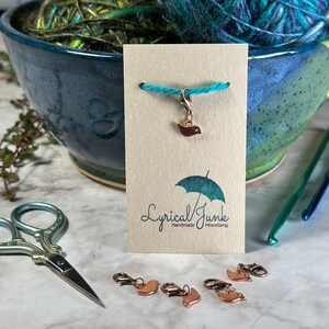 Peut inclure: Un ensemble de six marqueurs de points en or rose en forme d'oiseaux. Les marqueurs sont sur une surface blanche avec une paire de ciseaux bleu sarcelle, un crochet en bleu sarcelle et une carte brune avec le texte "Lyrical Junk Handmade Miscellany".