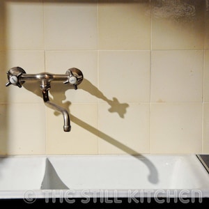 May include: A white porcelain double sink with a chrome faucet mounted on a tiled wall. The faucet has two handles and a spout. The sink is empty.