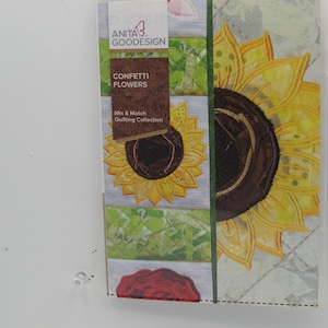 Puede incluir: Un primer plano de un panel de tela con un diseño de girasol. El panel es parte de una colección de acolchado llamada "Confetti Flowers" de Anita Goodesign. El panel es blanco con un borde verde y un centro marrón.