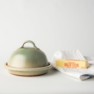 May include: A green ceramic butter dish with a lid and a stick of unsalted butter on a white linen tea towel. The butter wrapper says "Unsalted Butter Sweet Cream".