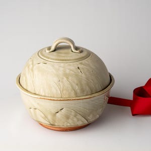 May include: A ceramic bowl with a lid, featuring a light green and beige color scheme with leaf-like patterns. The bowl has a rounded shape and a small handle on the lid. A red ribbon is visible on the side.