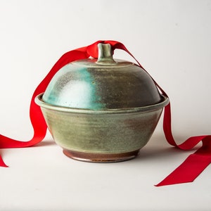 May include: A green ceramic bowl with a lid, tied with a red ribbon. The bowl has a rustic, handmade look.