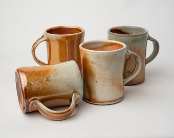May include: Four handmade ceramic mugs in shades of brown, tan, and cream. Each mug has a unique glaze and a curved handle. The mugs are arranged on a white surface, showcasing their individual shapes and textures.