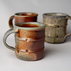 May include: Three handmade ceramic mugs with earthy tones. The mugs feature a cylindrical shape with a handle and decorative bands. The colors range from reddish-brown to tan and gray, with a textured finish. Each mug has a unique design.