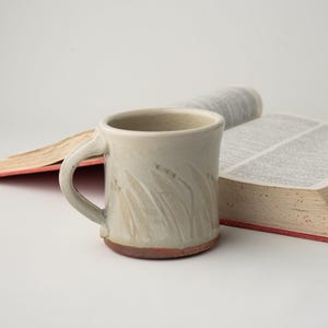 May include: A white ceramic mug with a textured design and a brown rim. The mug has a handle and is sitting next to a closed book.