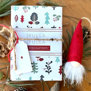 Op de afbeelding: Een stapel baksets verpakt in papier met een feestelijk boompatroon, vastgebonden met touw en een label. Een rood kabouterornament met een witte baard staat in de buurt. De sets zijn voorzien van het label "Rulla Pulla".
