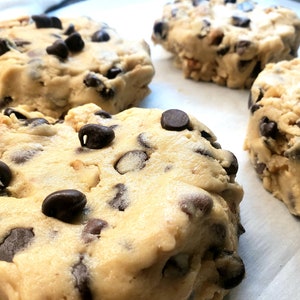 Pu&ograve; includere: Quattro palline rotonde di impasto per biscotti con gocce di cioccolato su una superficie di carta da forno bianca. L'impasto &egrave; di colore marrone chiaro e le gocce di cioccolato sono di colore marrone scuro.