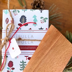 Op de afbeelding: Een rode en witte cadeaudoos met een feestelijk ontwerp met een kerstboom, sneeuwvlokken en bessen. De doos is vastgebonden met touw en een rood lint. De doos is gelabeld "Rulla Pulla" en bevat de tekst "Handgemaakt door jou! Premium bakset. Alle niet-bederfelijke ingrediënten. Volledige instructies inbegrepen."